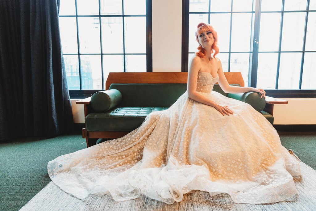 A wide shot of the bride sitting on the green leather couch in the hotel room with her dress spread out around her feet