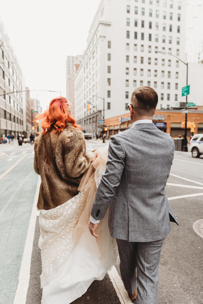 A photo of the bride and groom from behind walking to the car to take them to the ceremony and groom is carrying bride's dress