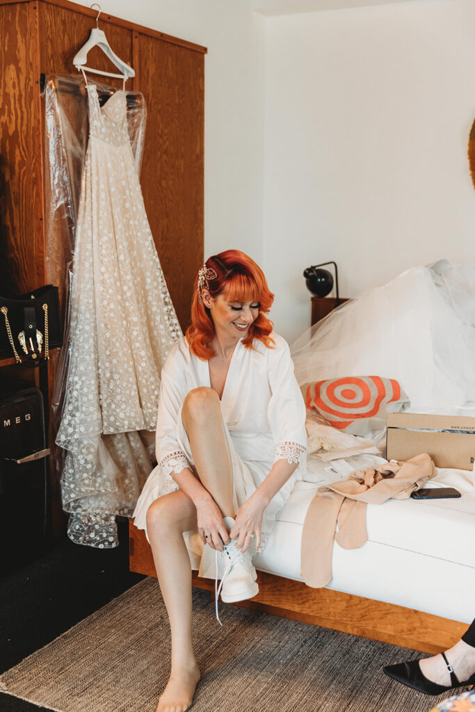 The bride getting ready and putting her shoes on in a hotel room with her dress hanging in the background