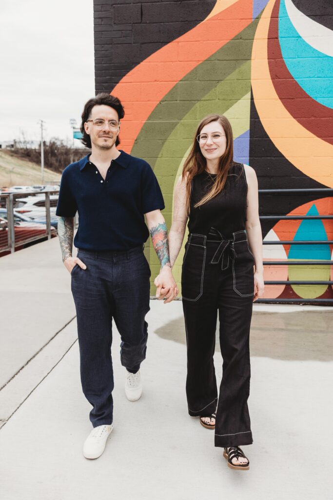 Couple in stylish denim coordinating outfits holding hands and walking in front of a bright and colorful mural that's outside of a brewery in downtown Baltimore. 