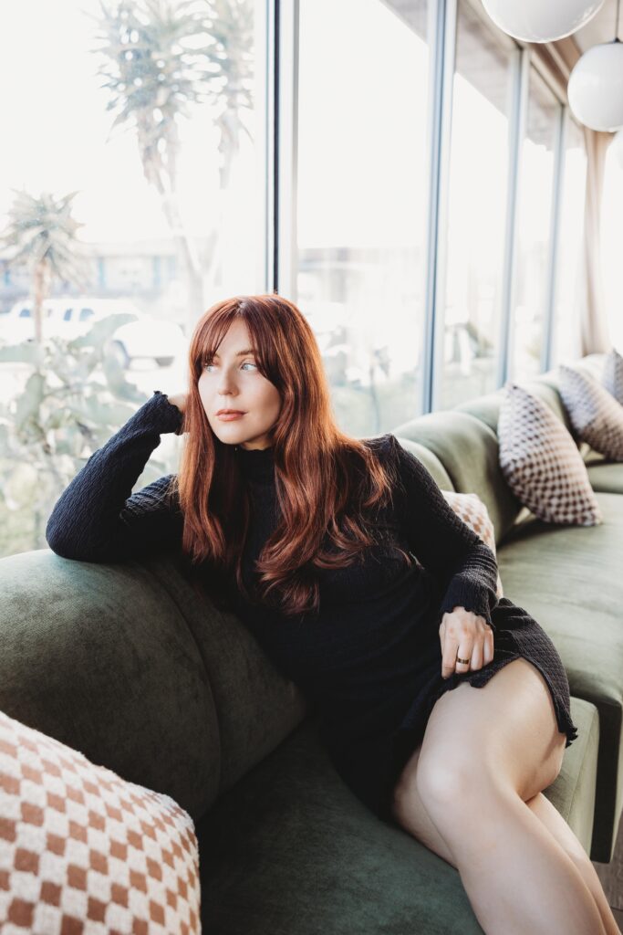 Red-headed woman is inside the Pearl Hotel now wearing a black long sleeve dress sitting on a dark green velvet bench with her legs crossed.
