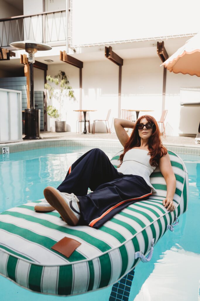 Red-headed woman wearing a bright white strapless peplum top and blue and orange athletic pants and Nike sneakers poses on a pool floatie in the pool in the hotel.