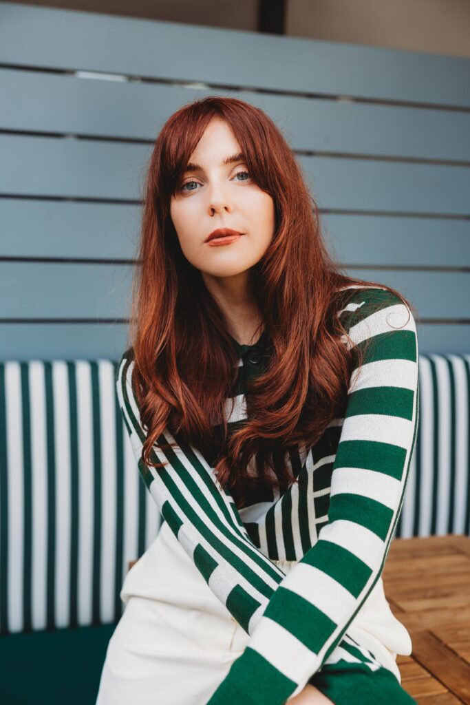 Red-headed woman with a striped white and green top sits on top of a patio table with her arms crossed and matches the bench behind her, also white and green stripes.